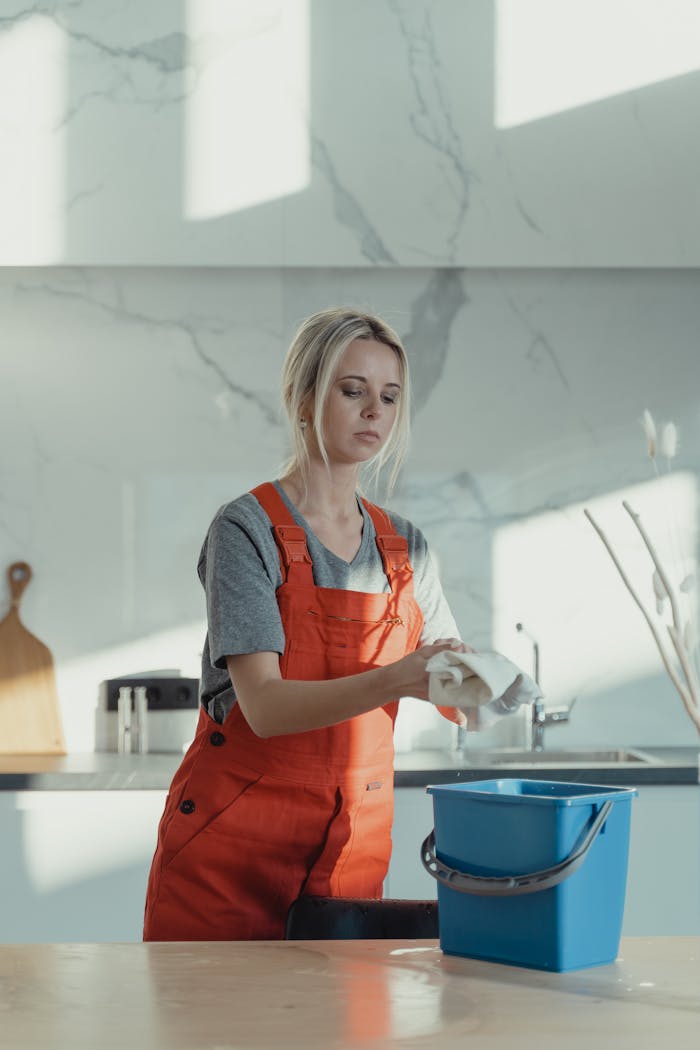 services-img A woman wearing orange overalls cleans a kitchen counter with a rag and pail in a modern kitchen.